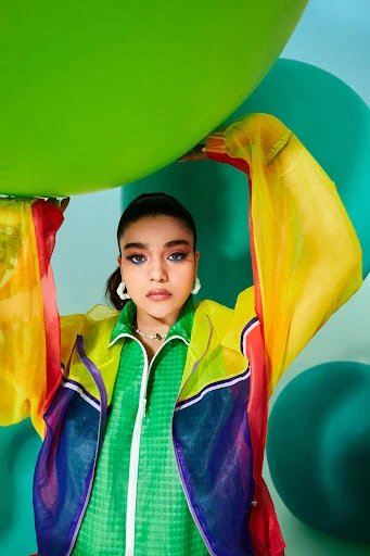 A young adult woman poses with a large green balloon, wearing a colorful sheer jacket, against a light blue backdrop with green balloons.