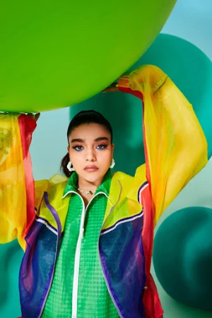 A young adult woman poses with a large green balloon, wearing a colorful sheer jacket, against a light blue backdrop with green balloons.