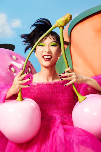 Energetic Asian woman in a pink tulle dress poses with oversized cherry props against a blue sky and cartoonish fruit background.