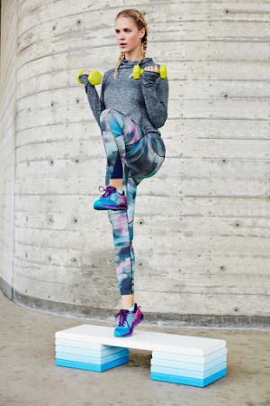 A woman in sportswear lifting a knee while holding dumbbells on a step in front of a concrete wall, exercising in daylight.
