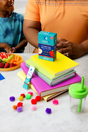 Hydration drink mix box on colorful books next to fruit platter and drinking cup with two humans present in soft indoor light.