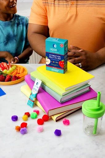 Hydration drink mix box on colorful books next to fruit platter and drinking cup with two humans present in soft indoor light.
