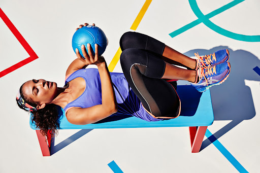 Athletic woman doing core exercises with a medicine ball on a workout bench in an indoor gym space with colorful geometric shapes.