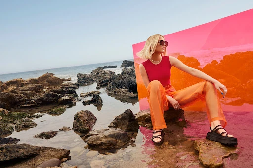 Young woman in red top and orange pants poses on rocks at a beach with a color panel, wearing sunglasses and platform sandals.