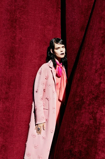 A fashionable woman poses indoors, wearing a pink coat, dress and tie against a dark red wall in a portrait-style editorial shot.