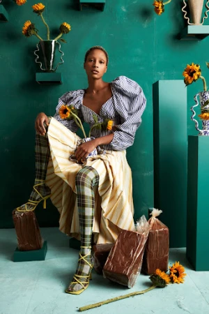 Young woman in patterned top and leggings, seated on bricks holding sunflowers against a green backdrop with flower-filled vases.