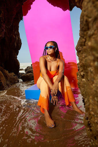 Young adult woman sitting on a blue cube by the water on the beach, framed by rocks and a magenta panel in the background.