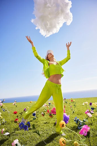 Woman in a green outfit poses outdoors under a cloud. Blue sky, green grass, and colorful flowers create a vibrant, eye-catching image.