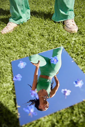 Woman in green activewear reflected in a square mirror on grass with scattered purple flowers, legs of person standing nearby.