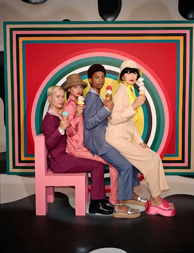 Four fashionably dressed people of diverse ages and ethnicities sit closely together on a pink chair eating ice cream cones indoors.
