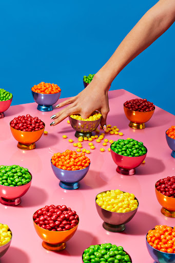 Overhead shot of hand reaching into a bowl of yellow candies, surrounded by bowls of red, green, and orange candies on pink table.