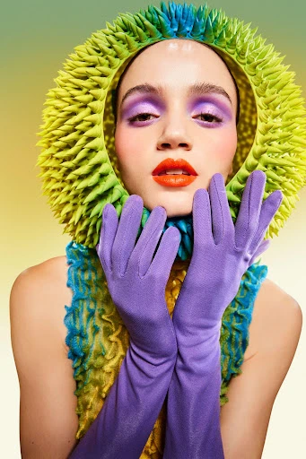 Woman in editorial fashion poses with spiky halo-like headdress, purple gloves, and vibrant eye makeup against a gradient background.