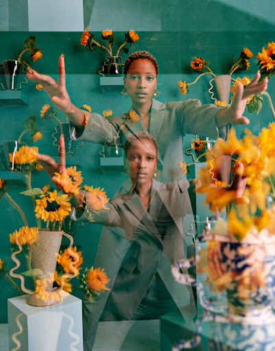 Surreal studio portrait of a black woman in an olive suit surrounded by sunflowers and geometric shapes, shot with soft, diffused lighting.