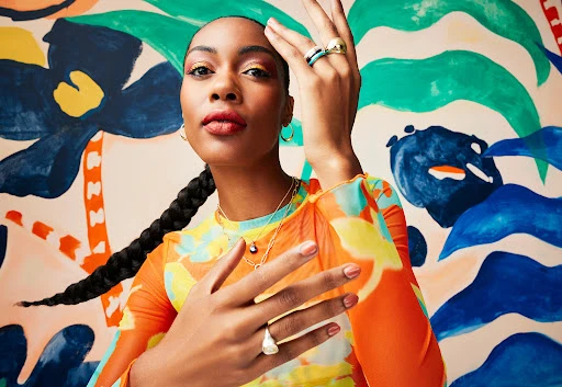 Portrait of a young Black woman with jewelry in a vibrant, artistic studio, featuring a colorful backdrop with abstract floral designs.