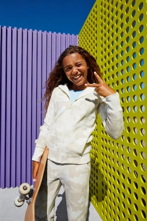 A smiling child in a patterned sweatsuit stands confidently with a skateboard against a colorful backdrop of yellow and purple panels.