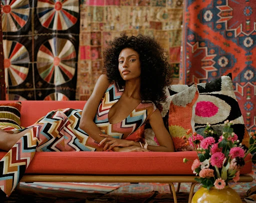 A striking fashion portrait features a model lounging on a vibrant coral sofa, framed by patterned textiles and a colorful flower arrangement.