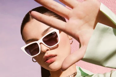 Close-up portrait of a woman wearing white sunglasses holding her hand up against a pink textured wall.
