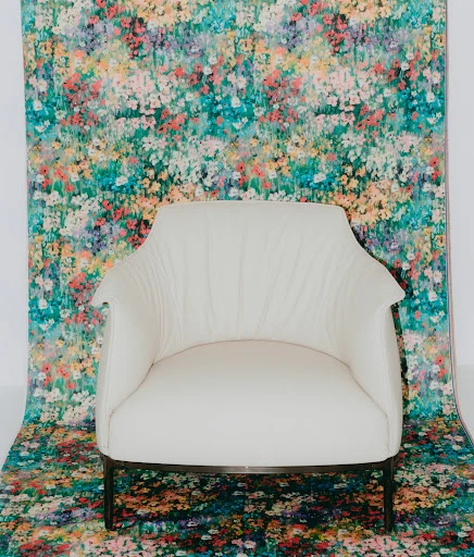 Studio shot of a white armchair on a thin metal frame, centered in the frame with a blurred floral backdrop and flooring.