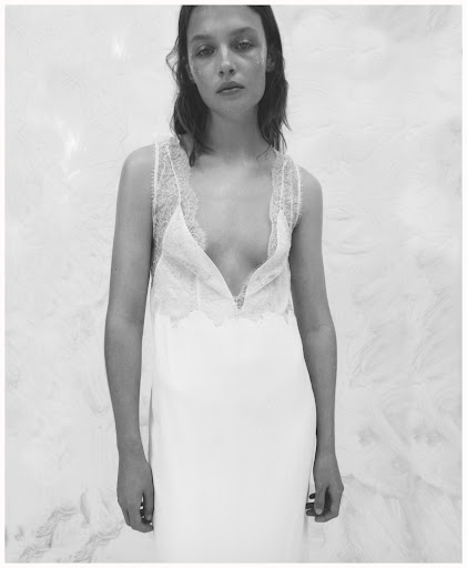 Monochrome portrait of a slender woman in a white, lace-trimmed dress, gazing at the camera with a neutral expression against a textured backdrop.