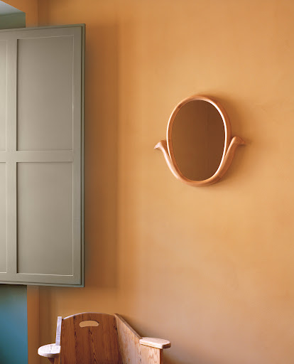 Interior shot of an oval mirror on a tan wall, complemented by a sage green cabinet and a light wood bench with rounded edges.