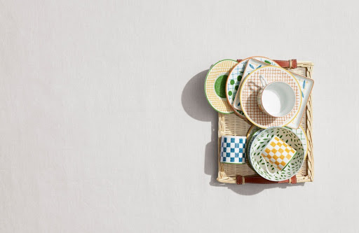 Overhead shot of assorted patterned plates, bowls, and mugs arranged on a woven tray against a pale background, studio lighting.
