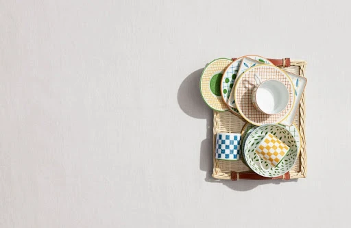 Overhead shot of assorted patterned plates, bowls, and mugs arranged on a woven tray against a pale background, studio lighting.