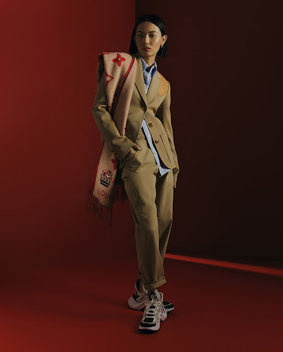 Asian woman in tan business suit poses indoors with patterned scarf and athletic shoes against a red wall in a fashion editorial.