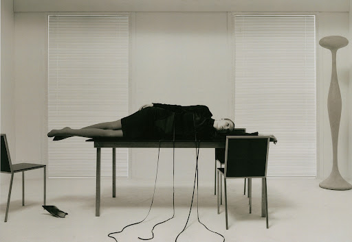 A woman lies on a table in a surreal, minimalist photo studio setting with chairs, a floor lamp, and cord cables draping downward.
