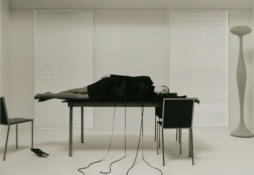 A woman lies on a table in a surreal, minimalist photo studio setting with chairs, a floor lamp, and cord cables draping downward.