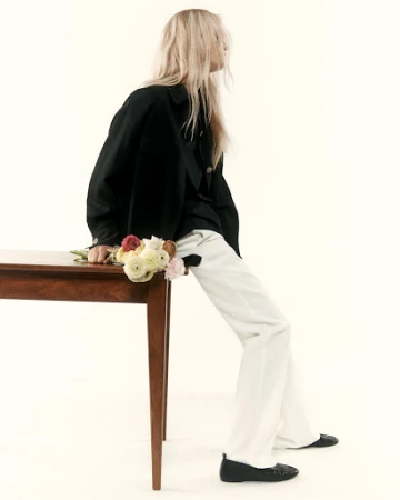 Blonde woman wearing a black jacket and white trousers leaning against a wooden table holding flowers