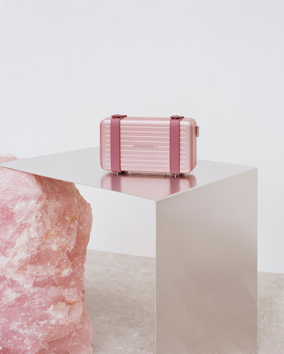 Small pink suitcase by Rimowa sits on a reflective surface next to a pink rock. A sleek and stylish still life.