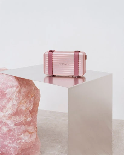 Small pink suitcase by Rimowa sits on a reflective surface next to a pink rock. A sleek and stylish still life.