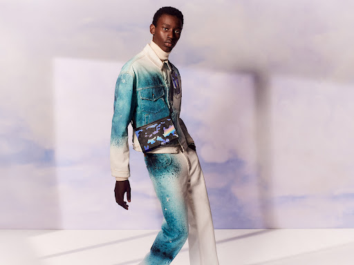 Dark-skinned adult male wearing a tie-dye denim jacket, matching pants, and crossbody bag poses in a brightly lit studio against a white backdrop.