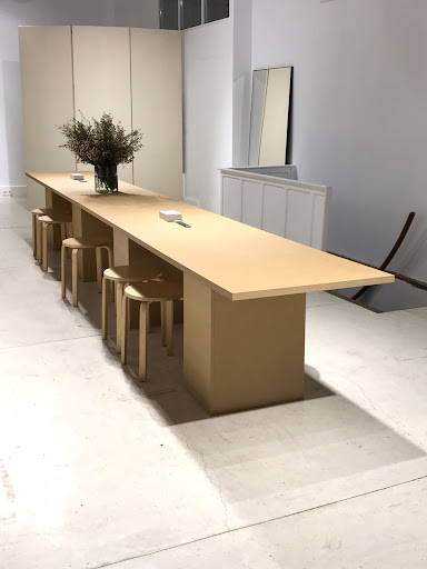 Minimalist dining space features a light wood table with matching stools, a vase, and minimalist decor against a neutral background.