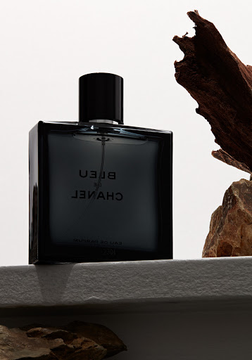 A square bottle of Bleu de Chanel perfume with brown wood shards on a textured white surface against a white background.