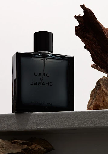 A square bottle of Bleu de Chanel perfume with brown wood shards on a textured white surface against a white background.