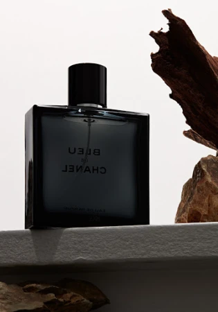 A square bottle of Bleu de Chanel perfume with brown wood shards on a textured white surface against a white background.