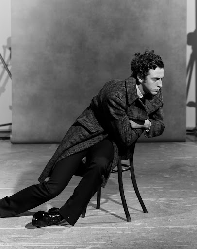 Black and white portrait of an adult male with curly hair sitting on a chair in a photo studio wearing a wool coat and dress shoes.