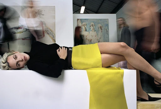 A young woman with blond hair lies on a white structure in a photo studio, wearing a black turtleneck and yellow skirt, with art in background.