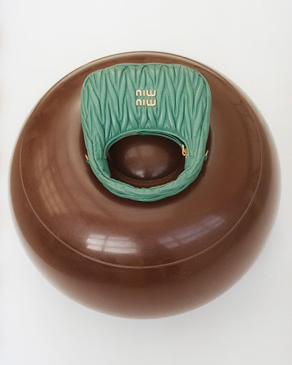 Green Miu Miu matelassé leather top handle bag on a round, shiny brown donut-shaped platform against a white backdrop in a studio setting.