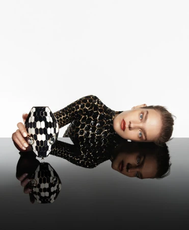 Woman in sequin top poses with a black and white patterned vase against a stark white and black reflective surface.