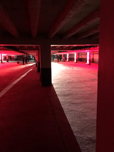 Underground parking garage with architectural columns and beams, lit with red lights, creating dramatic shadows and a stark, industrial aesthetic.