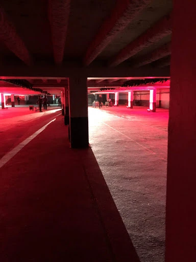 Underground parking garage with architectural columns and beams, lit with red lights, creating dramatic shadows and a stark, industrial aesthetic.
