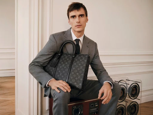 Adult male in a gray suit sits on a speaker, holding a Louis Vuitton handbag in an indoor setting, creating a high-fashion look.