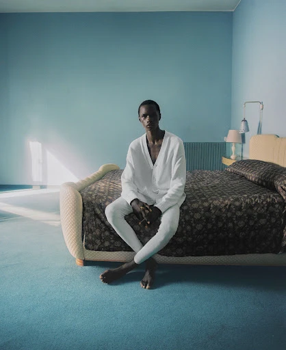 Young adult sits barefoot on bed in white outfit in minimalist bedroom with blue walls, matching carpet and brown floral bedding.