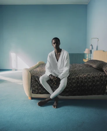 Young adult sits barefoot on bed in white outfit in minimalist bedroom with blue walls, matching carpet and brown floral bedding.