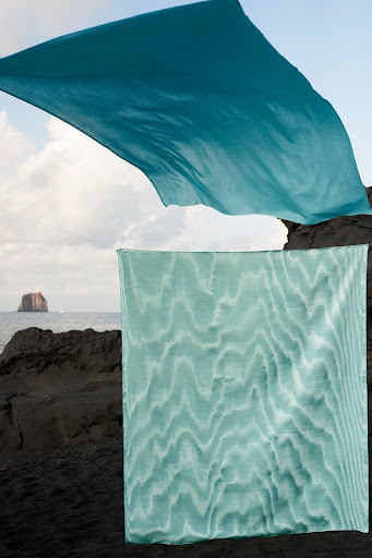 Two teal fabrics contrasting against black rocks and ocean, offering an abstract and serene beach scene with volcanic textures.