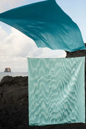 Two teal fabrics contrasting against black rocks and ocean, offering an abstract and serene beach scene with volcanic textures.
