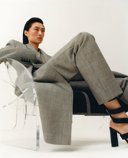 An Asian woman in a gray suit and black strappy heels reclines in a clear chair against a white backdrop in a fashion editorial studio shot.