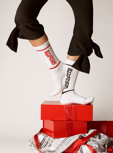Close-up shot of woman standing on glossy red gift boxes, wearing black pants tied at the ankles and branded white socks.